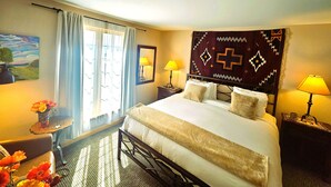 A hotel room with a bed, two bedside lamps, a window with curtains, a small table with a vase of flowers, and a painting on the wall. - Snakedance Condominiums (Taos Ski Valley)