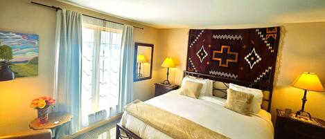 A hotel room with a bed, two bedside lamps, a window with curtains, a small table with a vase of flowers, and a painting on the wall.