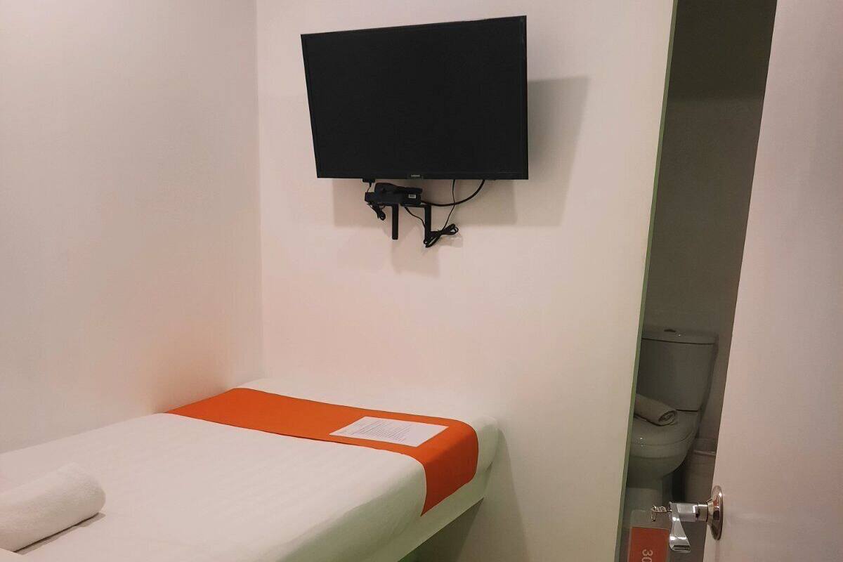 Un lit simple agrémenté d’une touche de couleur orange, un téléviseur à écran plat fixé au mur et une porte portant un numéro.