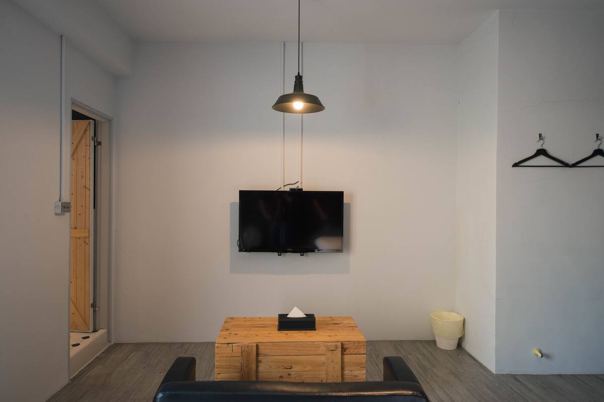 A room with a wooden chest, a television mounted on the wall, a hanging light fixture, and a door with a wooden frame.