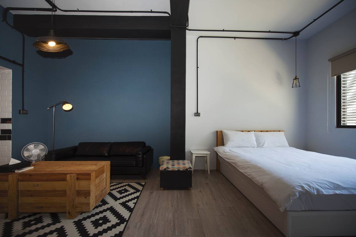 A compact bedroom with a bed, a small table, a sofa, a fan, and a patterned rug.