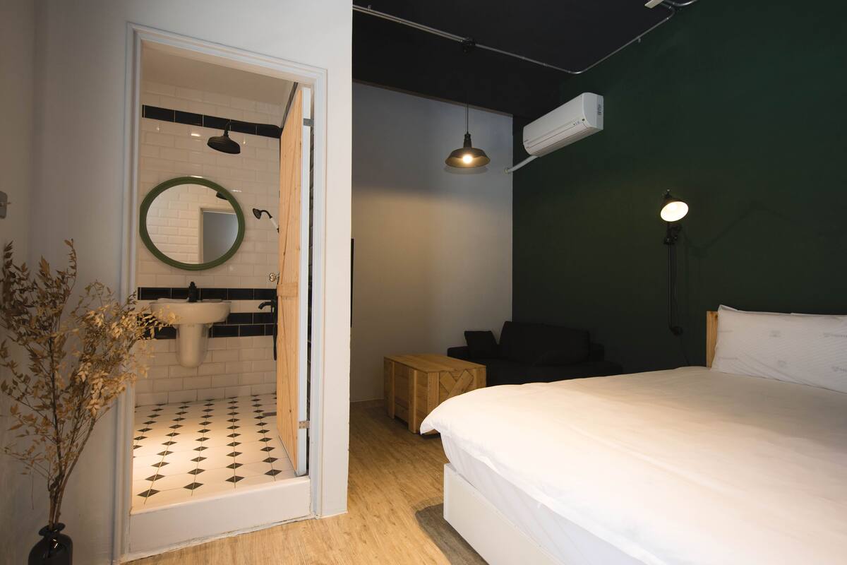 A bedroom with a bed, a bedside table, a mirror, a hanging light, and a view into the bathroom.