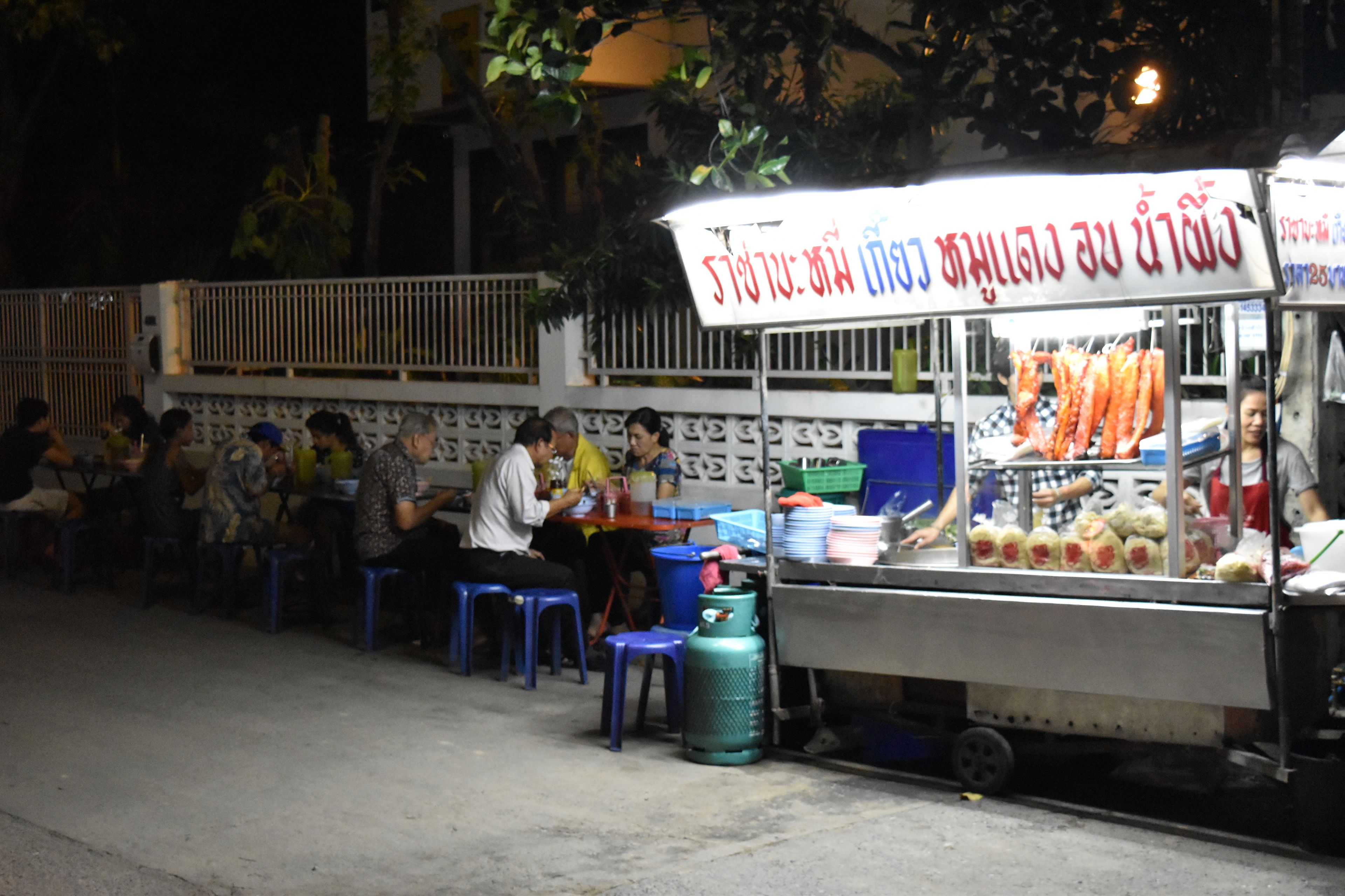 A street food stall with a sign in a foreign language, serving food to customers seated on stools.