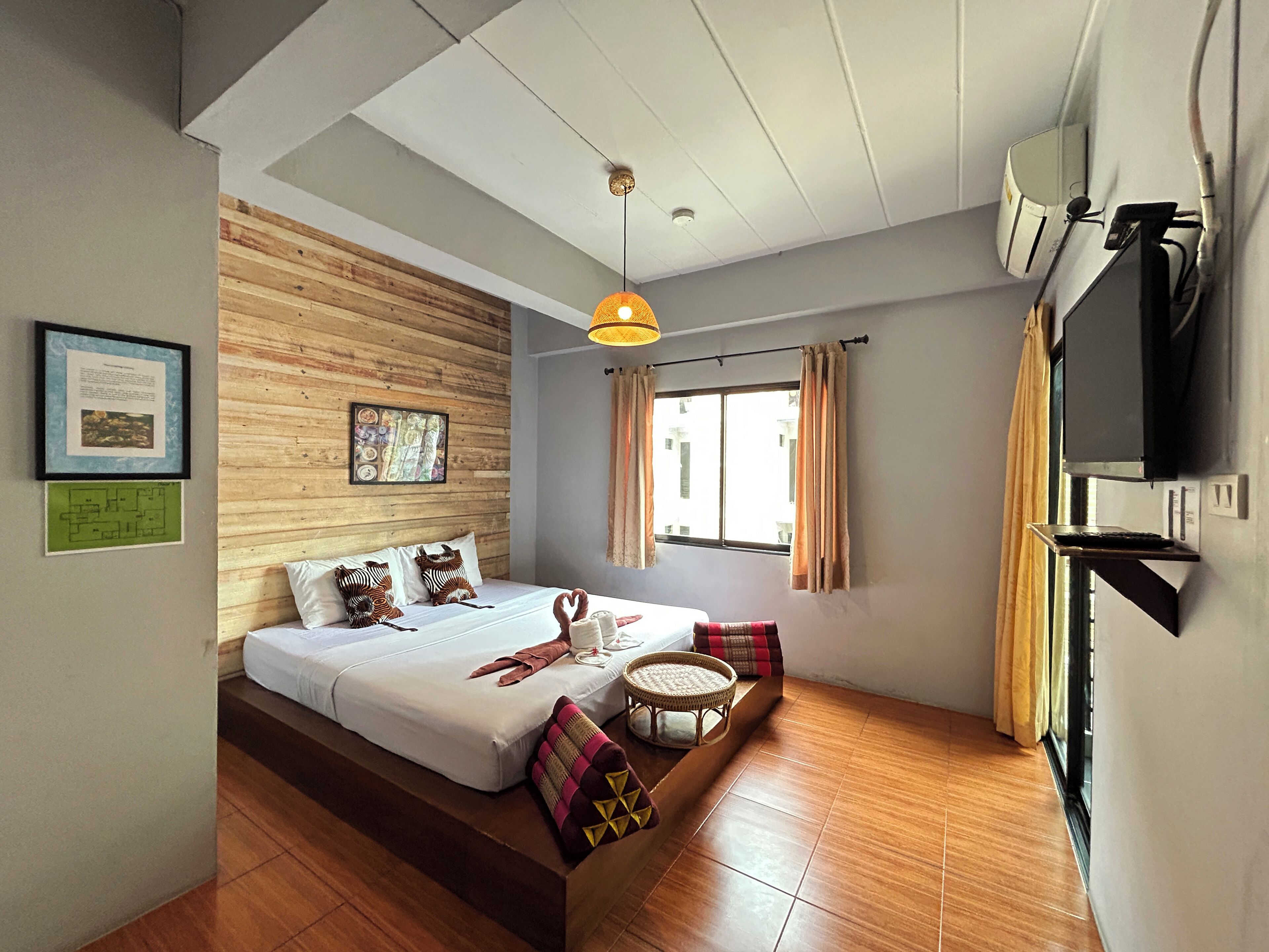Een moderne slaapkamer met een houten wand, een bed met veel kussens, een zithoekje, een raam met gordijnen en een tv aan de wand.