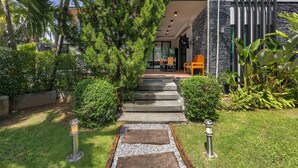 Un chemin menant à une maison moderne à la façade en pierre, entourée d’une végétation luxuriante et sous un ciel dégagé.
