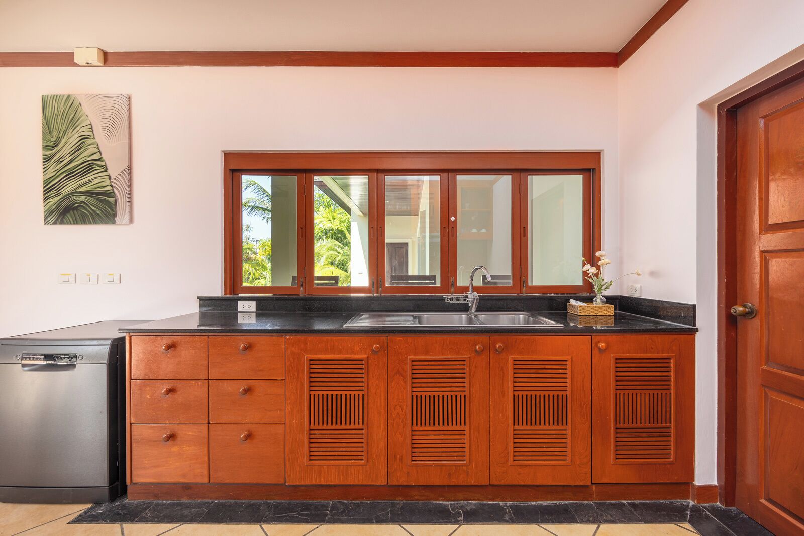 A kitchen with wooden cabinets, a stainless steel dishwasher, a black countertop, a sink, and a window with a view of palm trees.