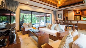 A spacious living area with a sofa set, a coffee table, and a TV. There is a dining area with a table and chairs. The room has large windows and a view of greenery outside. - Baan Bua Estate by Tropiclook (Rawai)