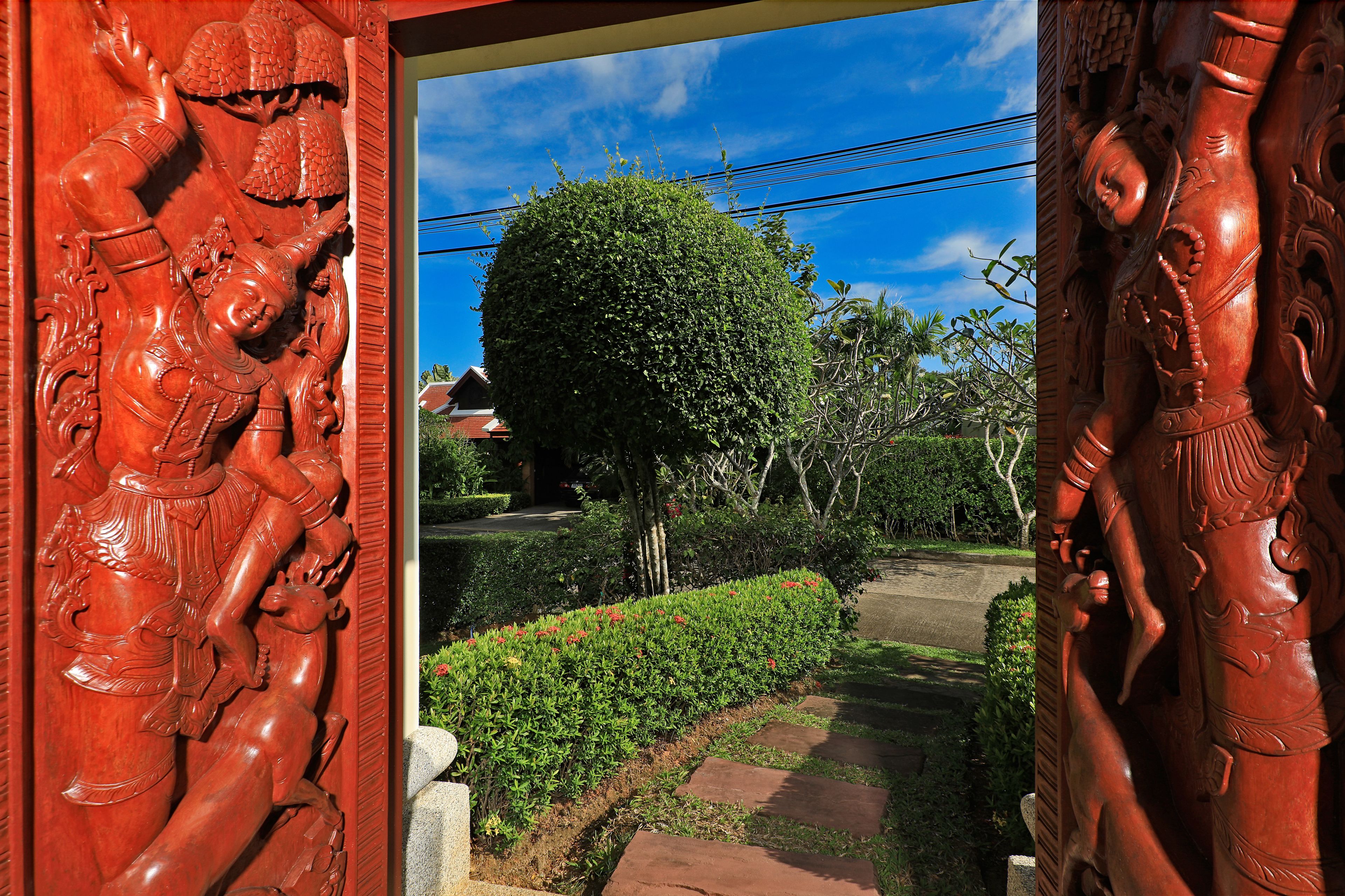 Una puerta de madera tallada con diseños intrincados, un jardín bien cuidado con un seto recortado y un pequeño árbol, y un cielo azul despejado.