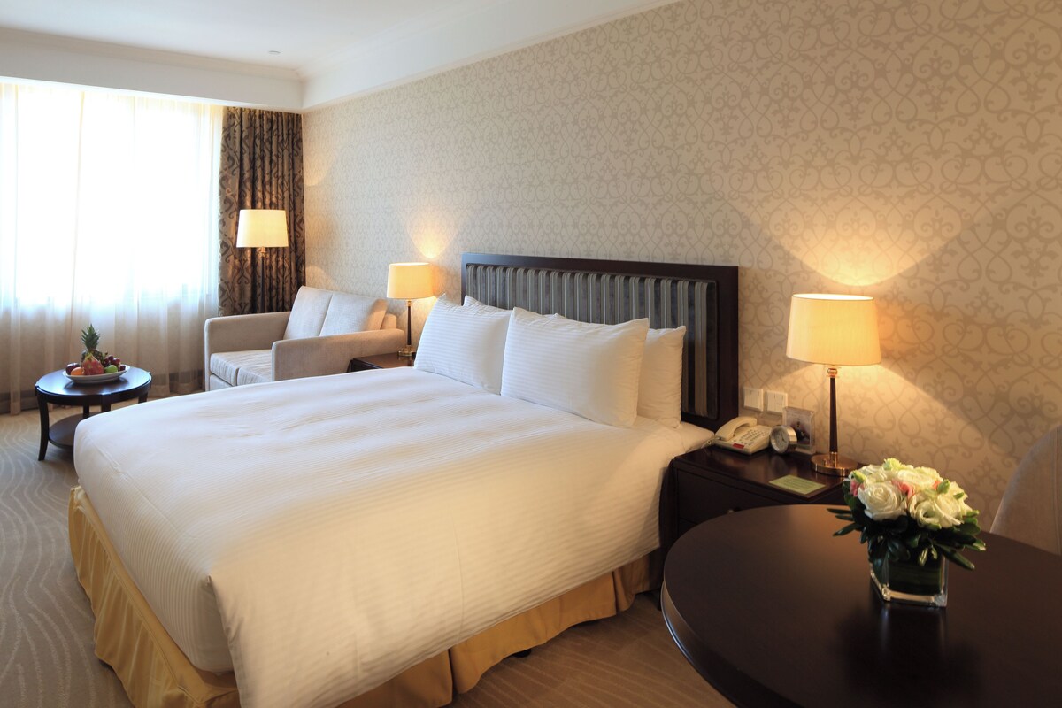 Habitación de hotel con una cama grande, dos mesitas de noche con lámparas, una pequeña mesa redonda con frutero y una silla.