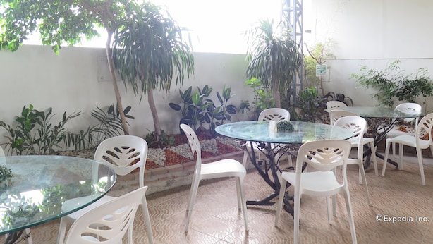 Un espacio de estar al aire libre techado con mesas de vidrio y sillas de plástico blanco, rodeado de plantas variadas y una pared blanca.