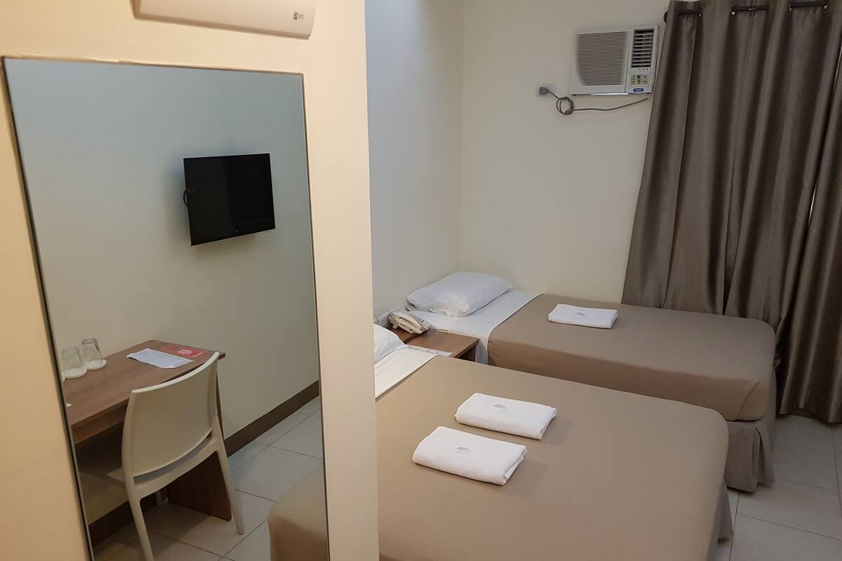 A hotel room with two beds, a desk, a chair, a flat-screen TV, an air conditioning unit, and a window with curtains.