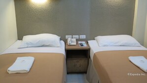 Habitación de hotel con dos camas, cada una con una almohada y una toalla doblada. Se observa una mesita de noche con un teléfono, un control remoto y una revista. La habitación tiene una pared con textura y un cabecero de madera.