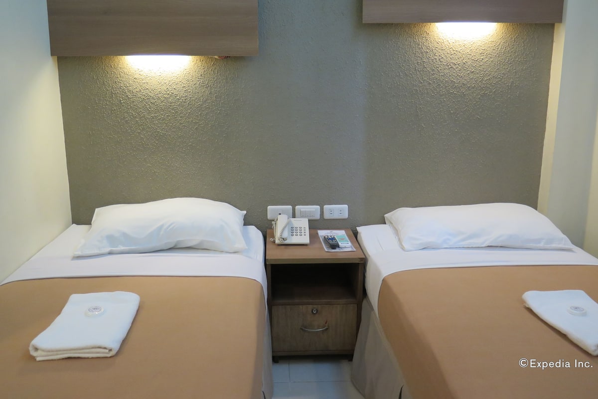A hotel room with two beds, each with a pillow and a folded towel. There is a bedside table with a phone, a remote control, and a magazine. The room has a textured wall and a wooden headboard.
