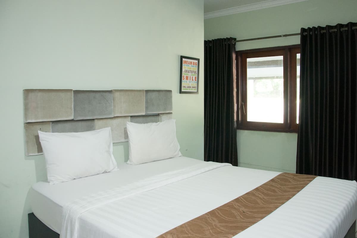A hotel room with a bed, a headboard with a plaque, a window with curtains, and a view of greenery outside.