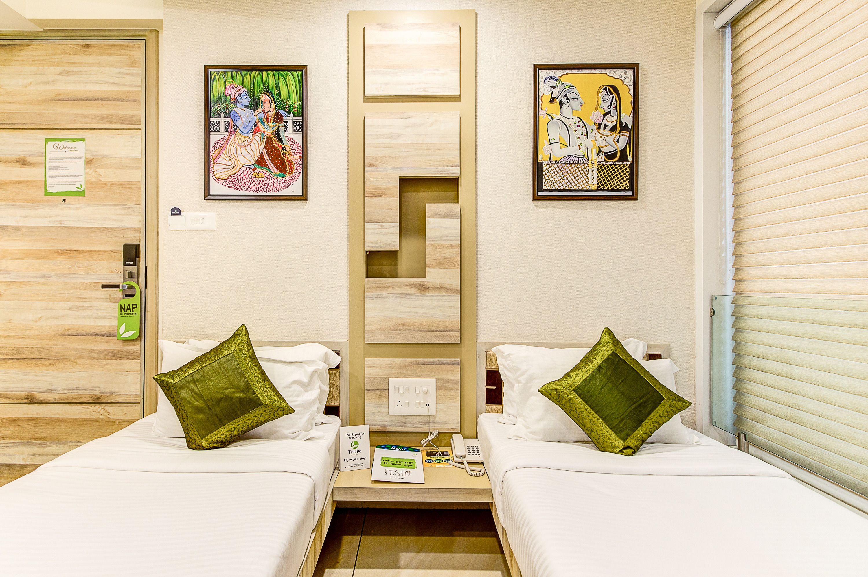 Habitación de hotel con dos camas, almohadas verdes, cabecera de madera, obras de arte en las paredes, un teléfono y un cartel que indica un servicio de SIESTA.