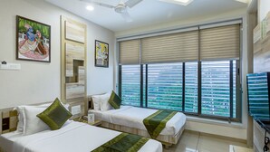 Une chambre d’hôtel avec deux lits, un ventilateur de plafond, une grande fenêtre avec des stores et des œuvres d’art accrochées aux murs.
