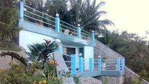 A multi-level building with a blue exterior and a stone base, surrounded by tropical vegetation. - High Steps Pension (Catmon)