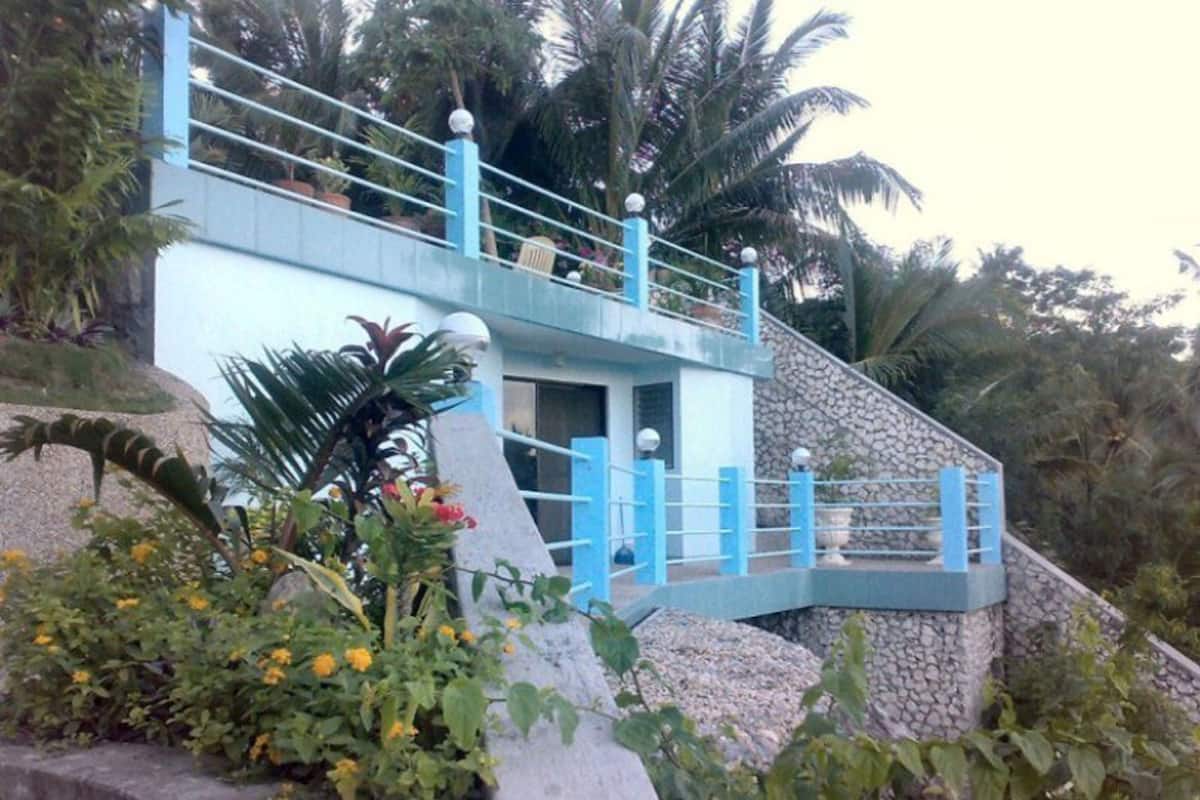 A multi-level building with a blue exterior and a stone base, surrounded by tropical vegetation.