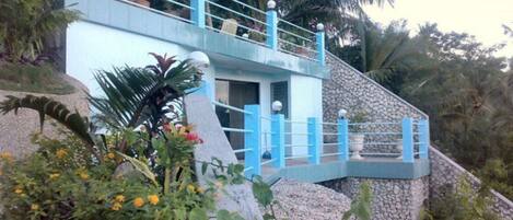 A multi-level building with a blue exterior and a stone base, surrounded by tropical vegetation.