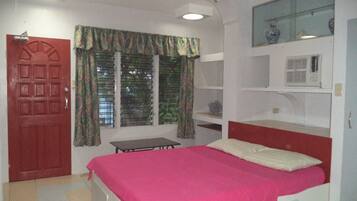 A bedroom with a bed, a window with curtains, a ceiling fan, and a shelf with decorative items.