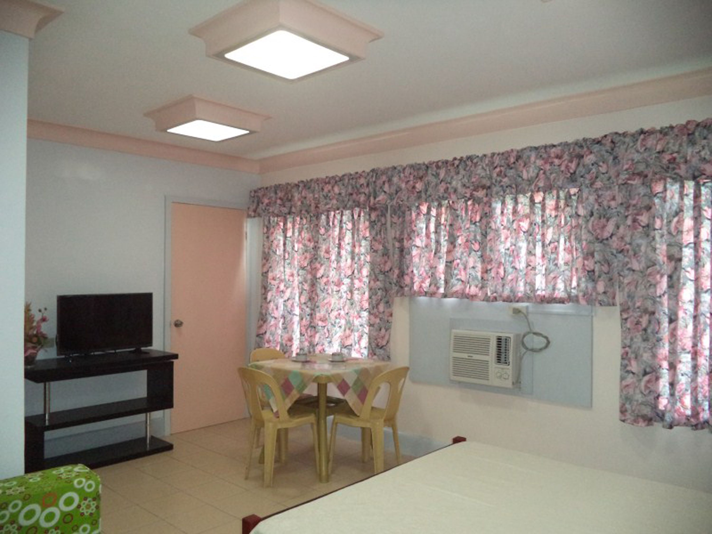A room with a bed, a desk with a television, a table with chairs, and floral curtains.