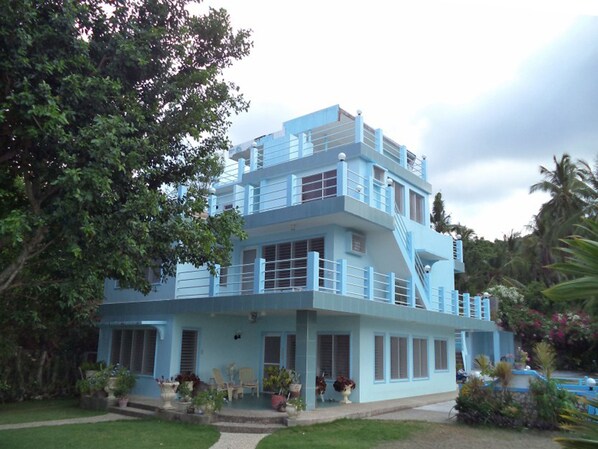 A multi-story building with a light blue exterior, a balcony on the upper floor, and a front porch with potted plants.