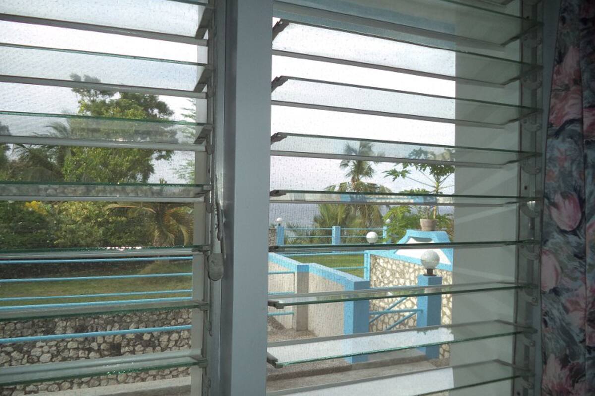 A view through a window with horizontal blinds, revealing a stone wall, a blue staircase, and some greenery outside.