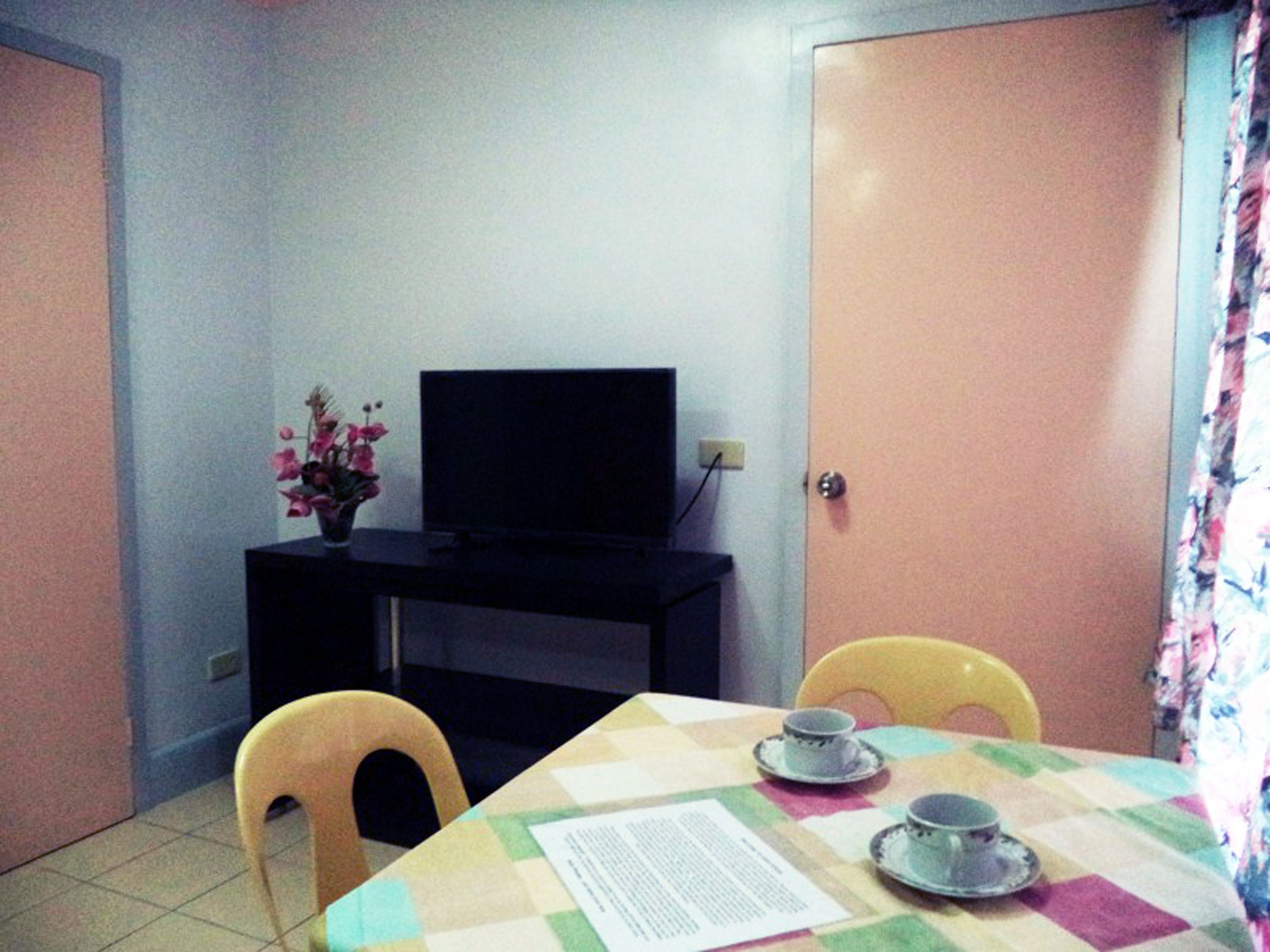 Una habitación pequeña con un televisor sobre un soporte negro, una mesa con mantel a cuadros, dos sillas y un jarrón con flores.