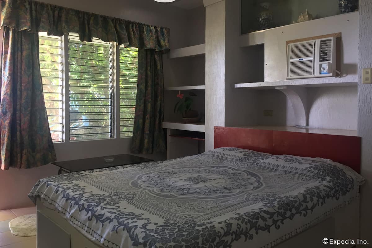 A bedroom with a bed, a window with blinds, a shelf with decorative items, and a ceiling light.