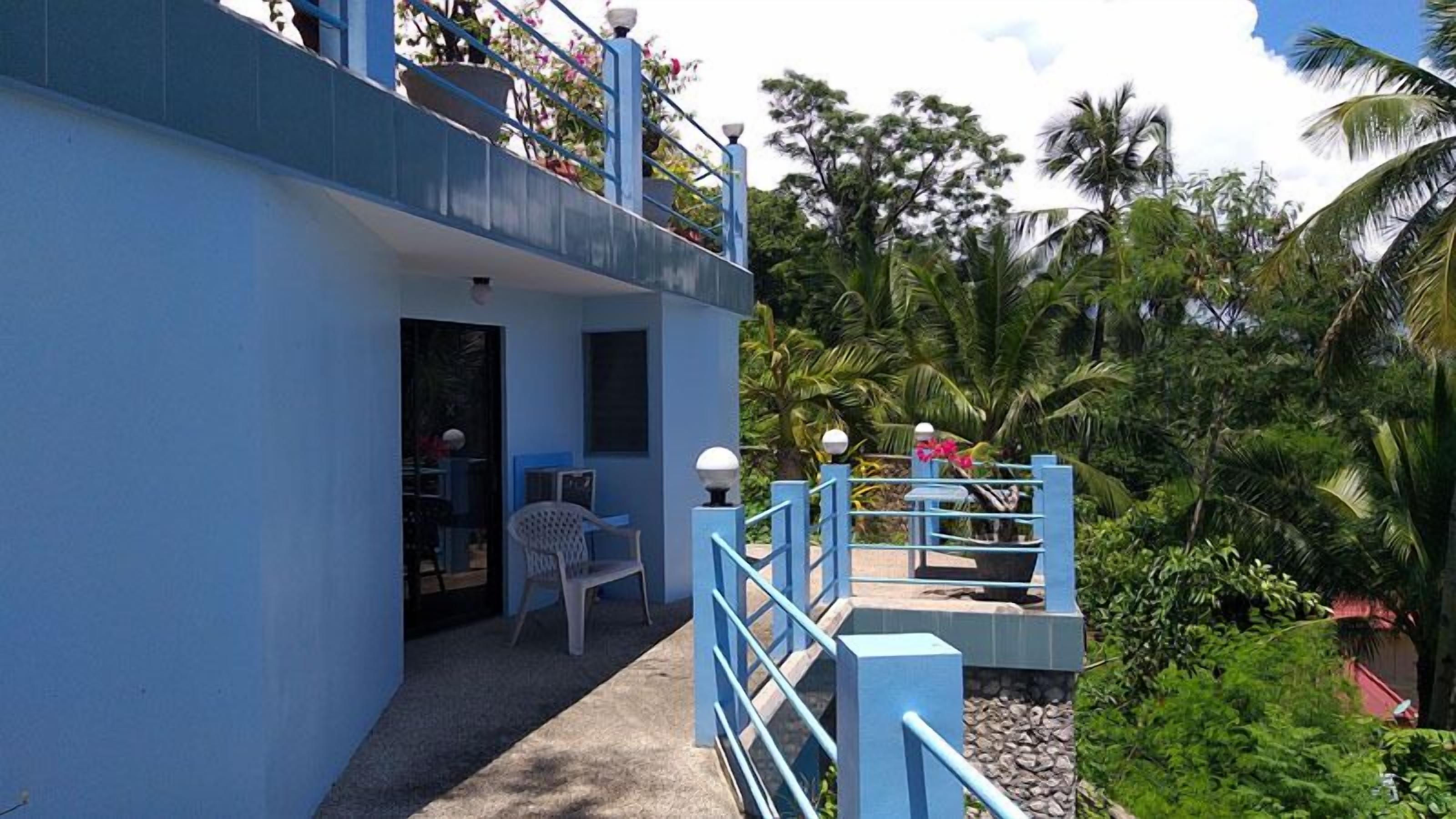 A two-story building with a balcony, a small outdoor seating area, and a view of tropical vegetation.