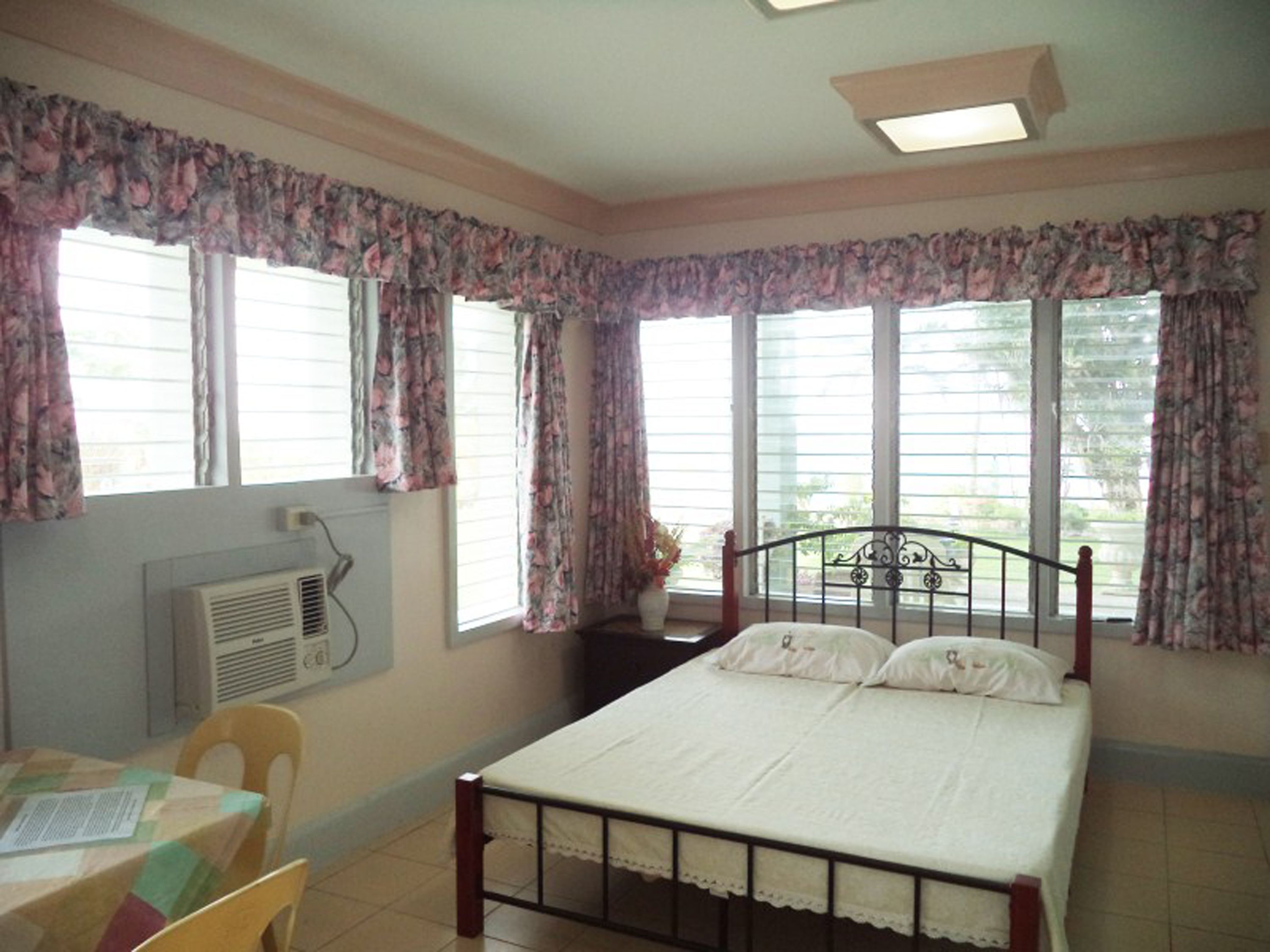 Un dormitorio con cama, escritorio y silla. Se observan ventanas con cortinas, un equipo de aire acondicionado y una lámpara en el techo.