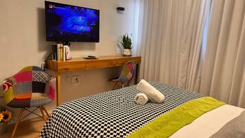 A hotel room with a bed, a desk, a chair, a TV, and a potted plant.