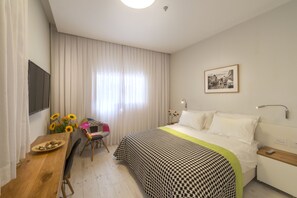 A hotel room with a bed, a desk with a chair, a window with curtains, a wall-mounted TV, and a framed picture on the wall. - Dizengoff 208 Hotel (Tel Aviv)