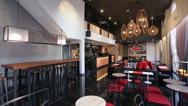 Zona de bar moderna con taburetes altos, una barra larga y un área de asientos con sillas rojas y negras.