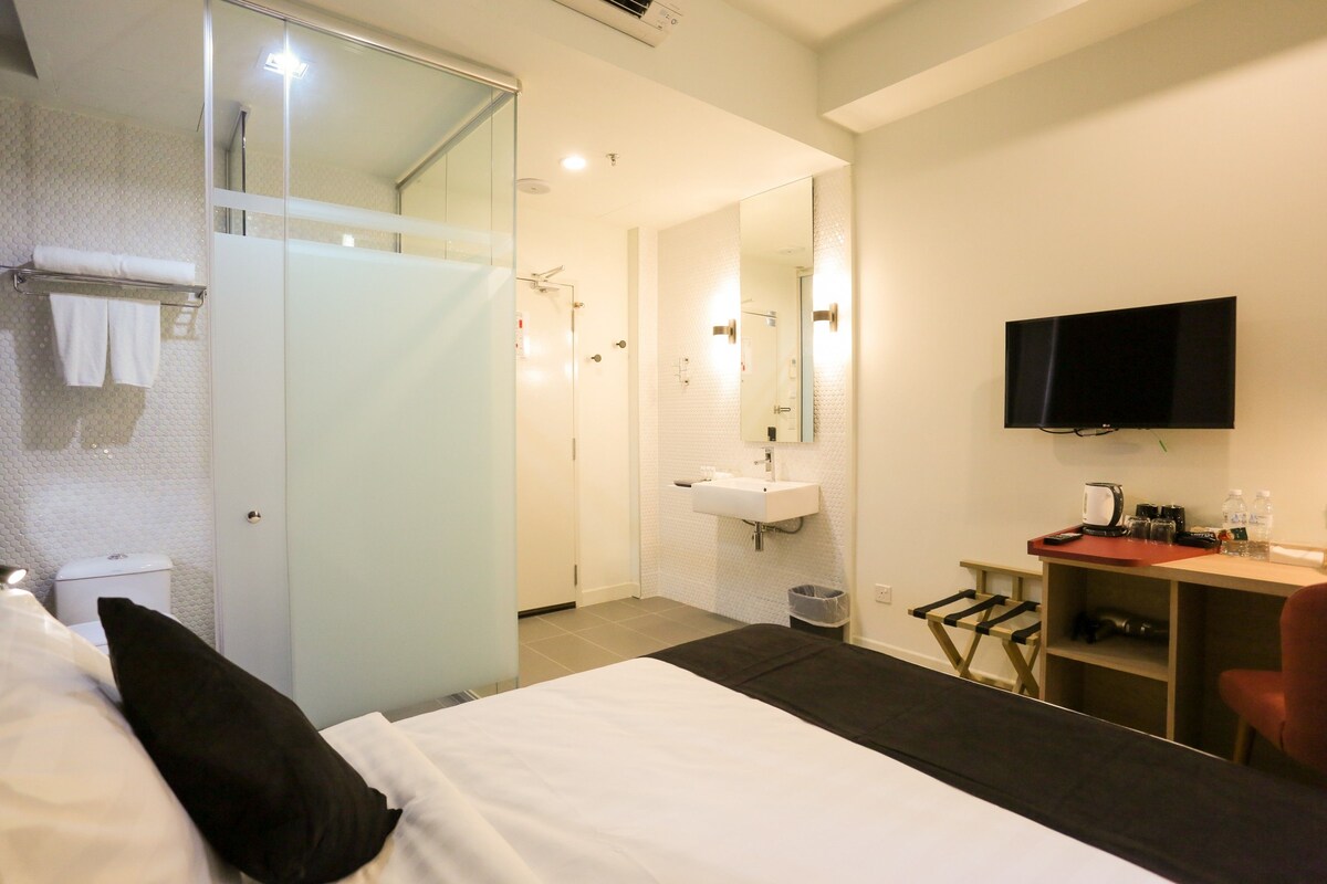A hotel room with a bed, a TV, a desk with a chair, a bathroom with a shower, and a sink.