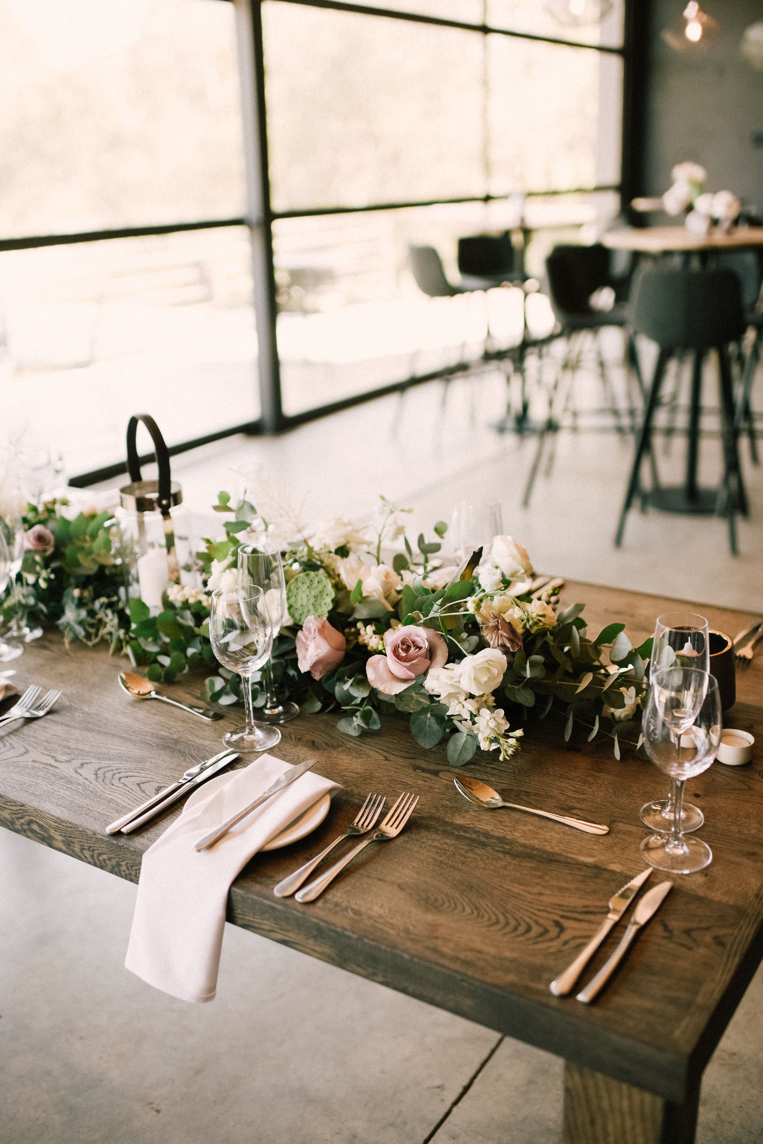 A wooden table set with white napkins, silverware, and wine glasses, adorned with a floral centerpiece.