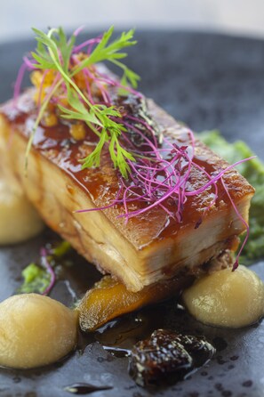 A dish with a glazed pork loin, garnished with purple microgreens and a yellow puree, served on a dark plate. - Cradle Boutique Hotel (Muldersdrift)