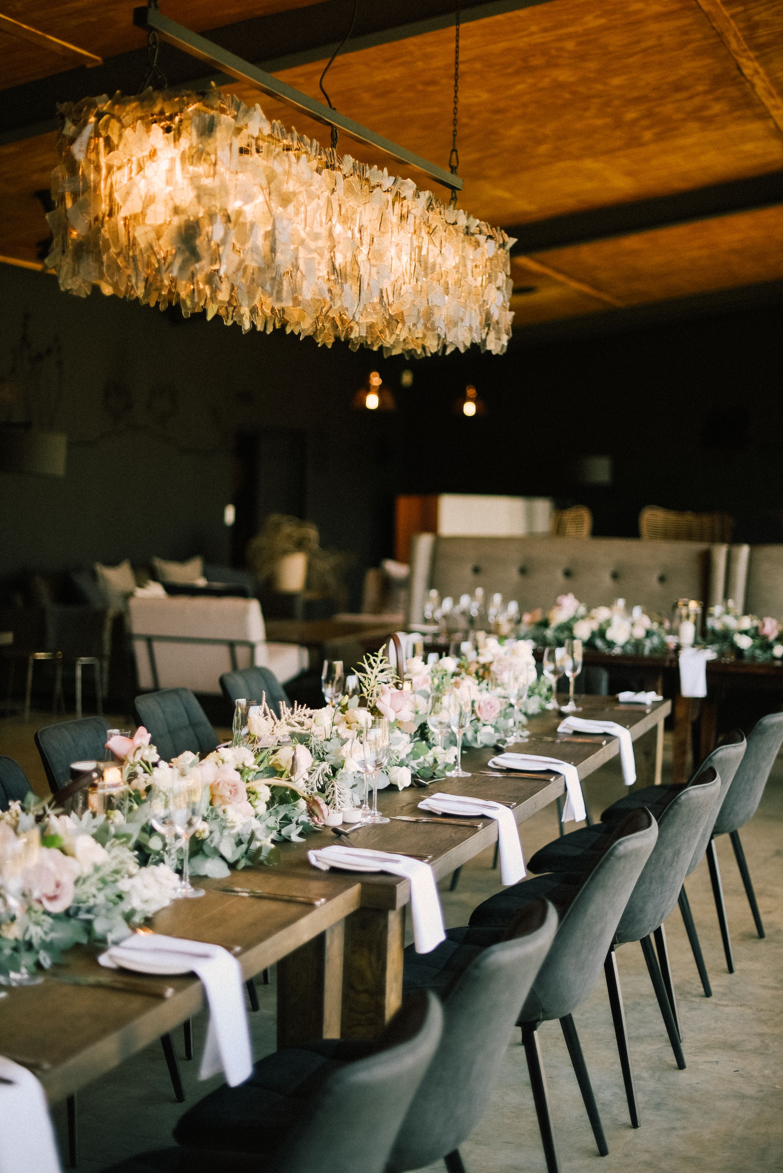 Una larga mesa de comedor con sillas, una araña y centros de mesa florales.