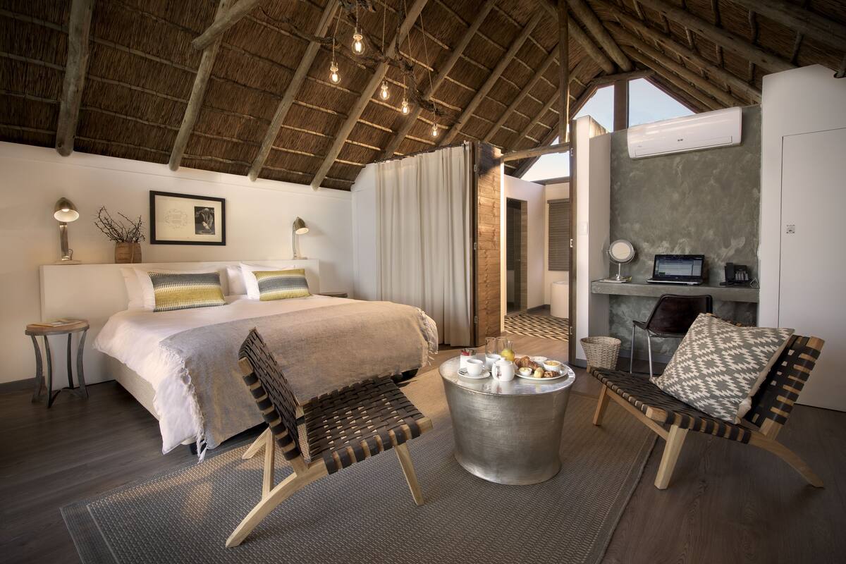 A hotel room with a bed, two chairs, a small table, a desk, and an air conditioning unit.