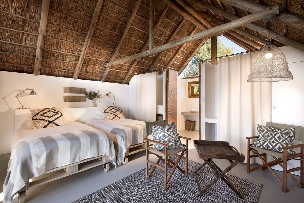 A cozy bedroom with a bed, two chairs, a small table, a hanging light fixture, and a window with curtains.