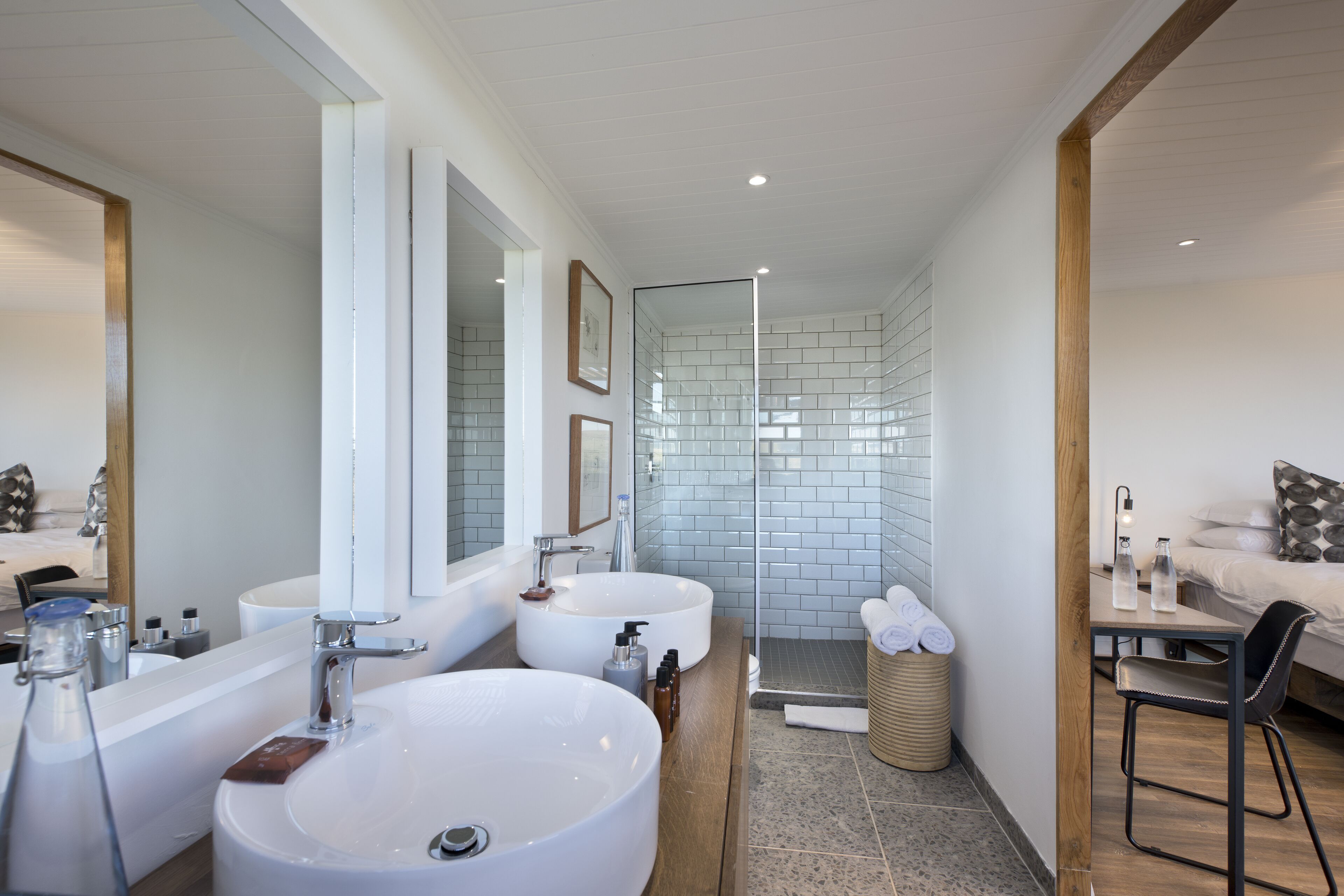 Une salle de bain moderne avec deux grands lavabos, une douche à l’italienne en verre et une petite table avec une chaise.