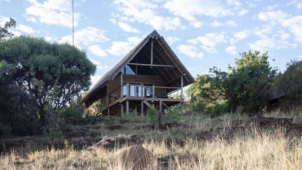 A wooden cabin with a triangular roof, built on stilts, surrounded by tall grass and trees. - Cradle Boutique Hotel (Muldersdrift)