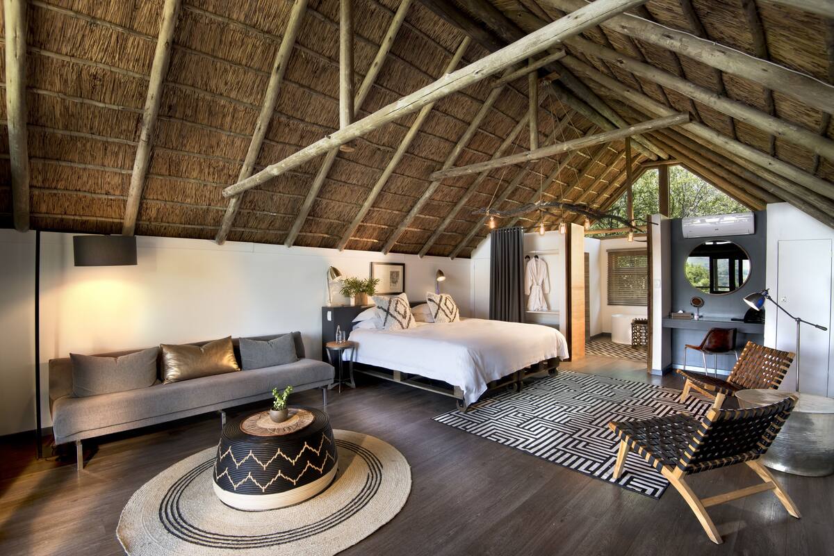 A bedroom with a bed, a sofa, a coffee table, and a chair. There is a ceiling with wooden beams and a thatched roof.