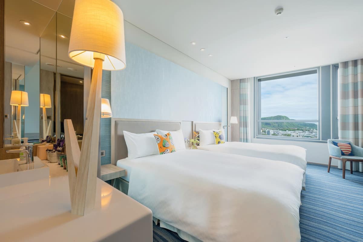Habitación de hotel con dos camas, un ventanal con vistas, un baño con lavamanos y un espejo.