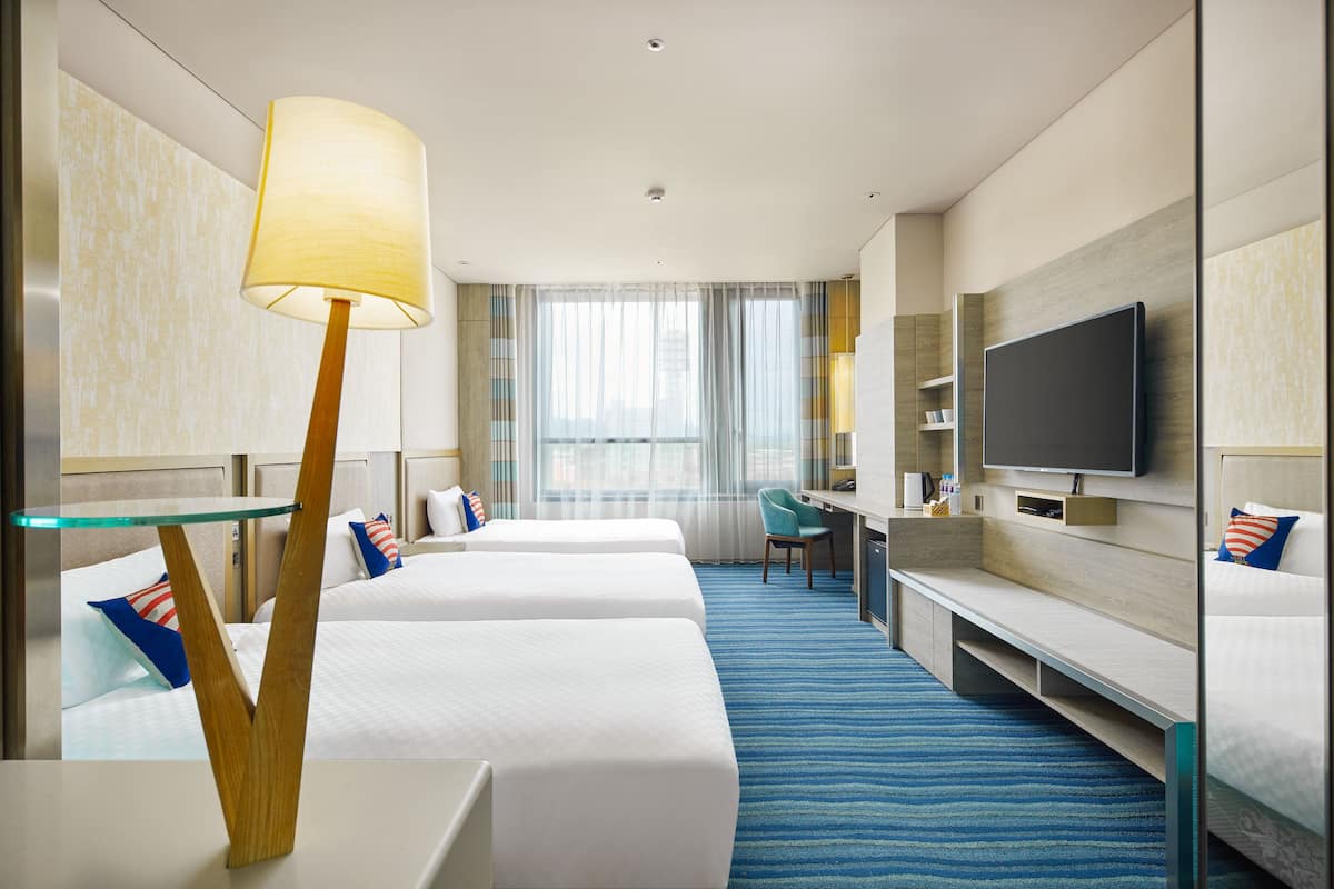 Habitación de hotel con tres camas, un escritorio, un televisor y un ventanal con cortinas.