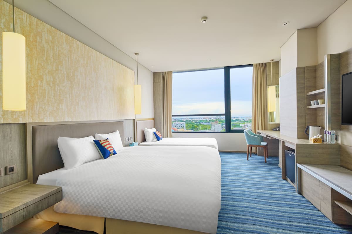 Habitación de hotel con dos camas, un escritorio, una silla, un televisor y un ventanal grande con vistas a la ciudad.