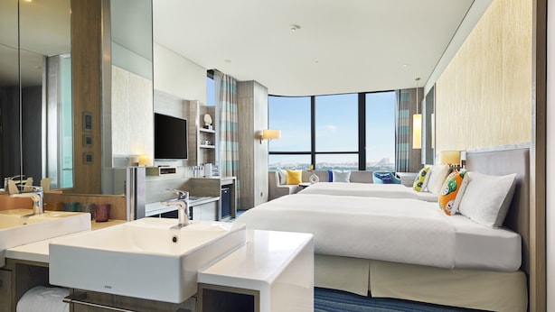 Une chambre d’hôtel moderne dotée d’un grand lit, d’un canapé séparé, d’une salle de bain avec un lavabo et un miroir, et offrant une vue sur la ville grâce à de grandes fenêtres.