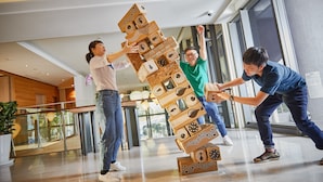Un groupe de personnes joue avec une grande structure en bois à plusieurs niveaux, dans un espace intérieur vaste et moderne, doté de grandes fenêtres et d’un plancher ciré.