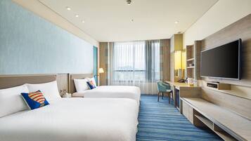 A hotel room with two beds, a desk, a TV, and a large window with curtains.