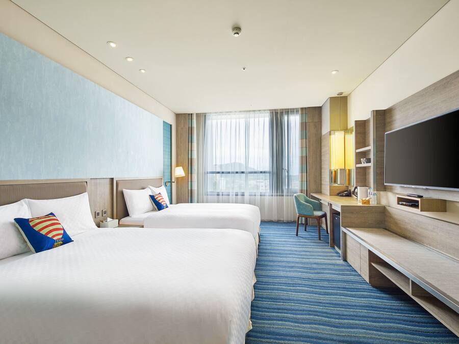 A hotel room with two beds, a desk, a TV, and a large window with curtains.